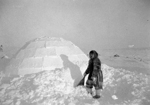 Black and white photograph of a man wearing a fur parka, standing with a wooden shovel in hand beside an igloo surrounded by snow