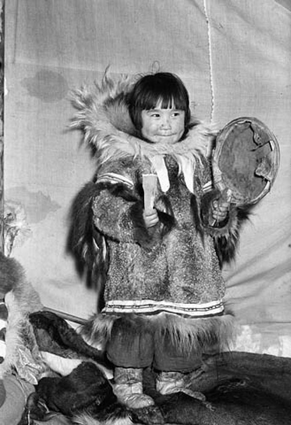 Black and white photograph of a young girl dressed in elaborate winter clothing, holding a drum