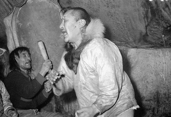 Black and white photograph of two men inside a tent; one is playing a drum, while the other is singing and dancing.