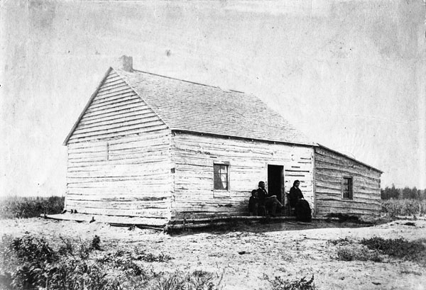 Black and white photograph of a wooden prairie house with a sloped roof and three people sitting outside on a porch