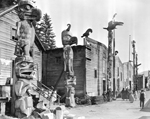 Black and white photograph of a street scene with large wooden houses and totem poles lining the wooden walkway in front of each house