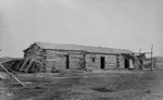 Black and white photograph of a long, single-storey log home with several doors, and tools and implements surrounding the building