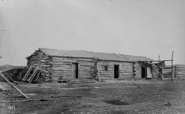 Black and white photograph of a long, single-storey log home with several doors, and tools and implements surrounding the building